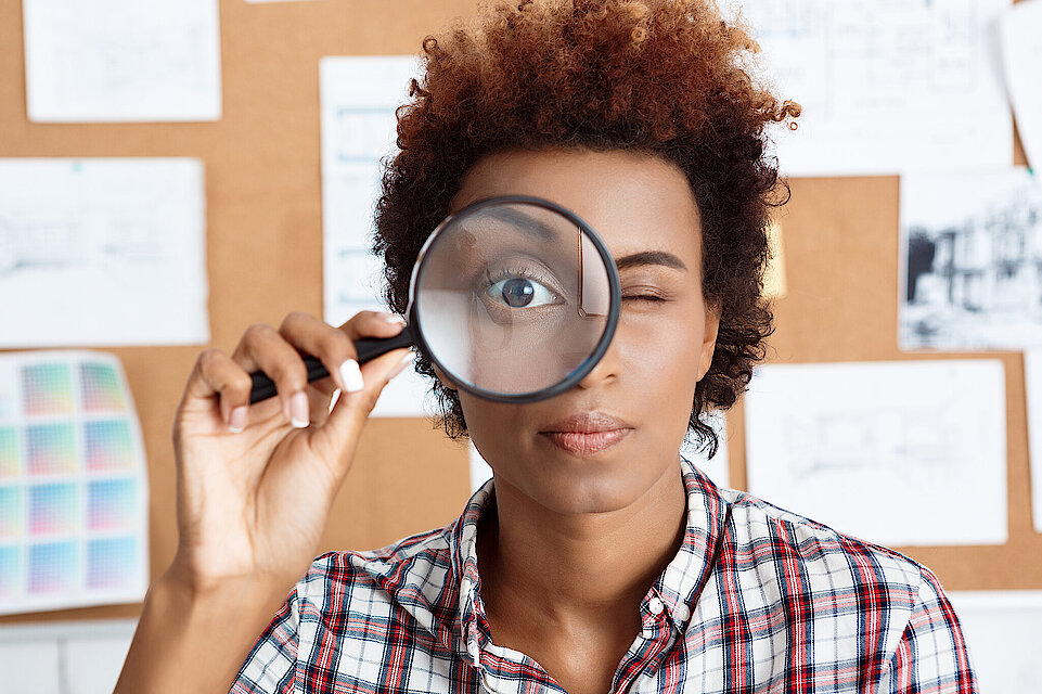 Young beautiful african worker holding magnifier infront of eye. Office background.